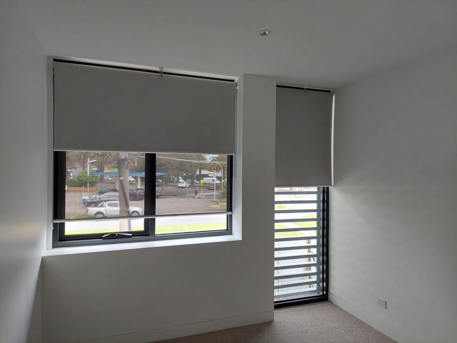 Grey roller blinds fitted to twin windows in a Melbourne bedroom, with plantation shutters visible on an adjoining window.