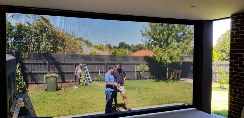 View through a retractable mesh door screen of two people talking in a backyard.